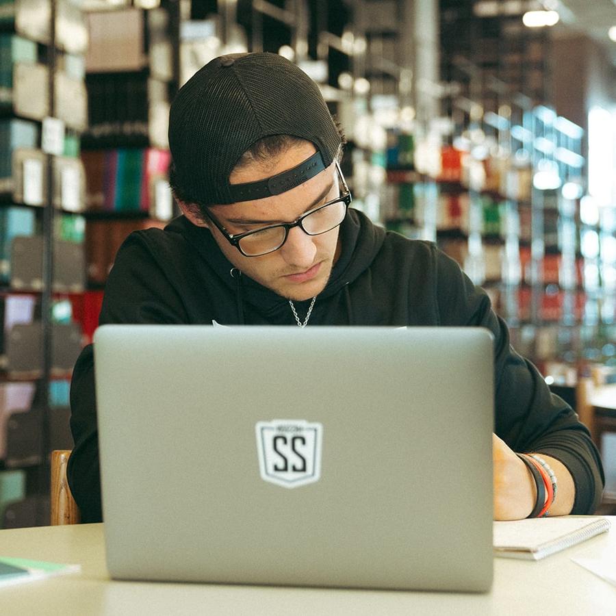 Male student at laptop in library with books in background.